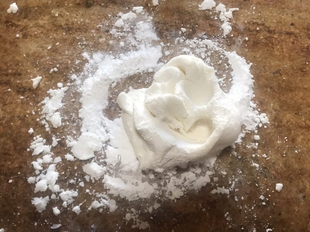 A small mound of homemade white gum paste sits on a lightly dusted surface with powdered sugar scattered around.