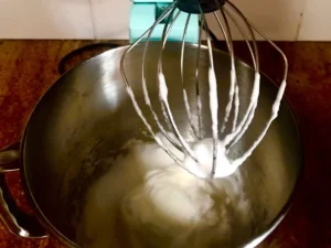 Aqua KitchenAid stand mixer with stiffly whipped egg whites in a stainless steel bowl on a kitchen counter.