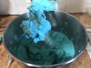 Bright turquoise buttercream frosting in a stand mixer bowl, with some of the frosting clinging to the paddle attachment above.