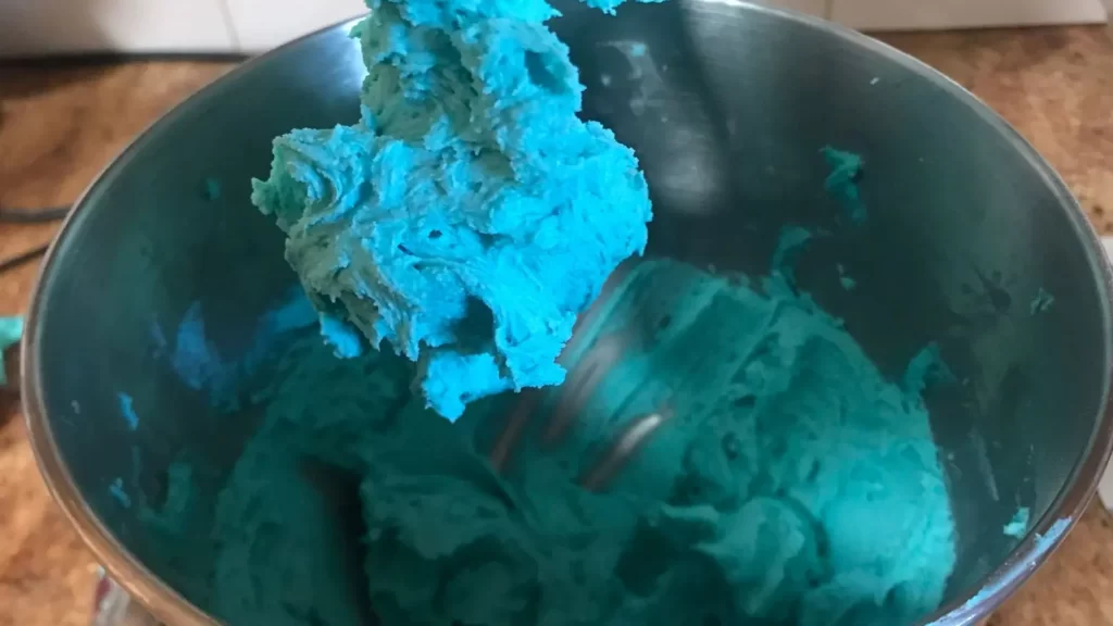 Bright turquoise buttercream frosting in a stand mixer bowl, with some of the frosting clinging to the paddle attachment above.