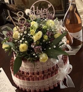 Two-tier money cake decorated with yellow roses, purple flowers, lace ribbon, and a Happy Birthday topper, with a bottle of bubbly beside it.