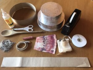 Materials laid out for assembling a two-tiered money cake, including cake boards, cardboard strips, glue, tape, paper clips, ribbon, R50 South African banknotes, and crafting tools.