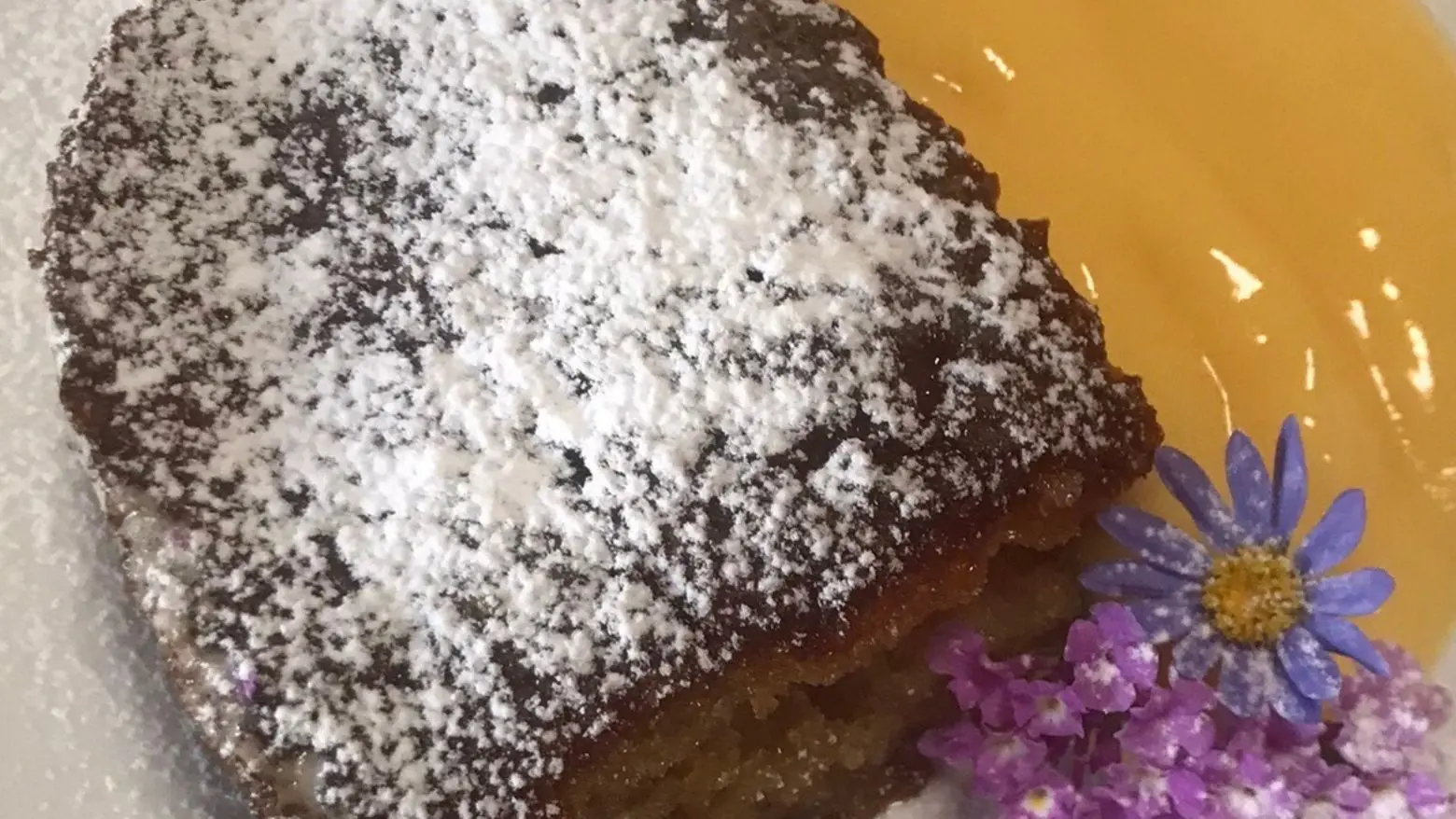 A slice of golden brown Malva Pudding dusted with powdered sugar, served with a generous portion of smooth custard on a white plate, garnished with delicate purple and blue flowers.