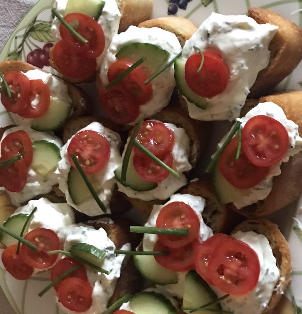 A plate of crispy baguette slices topped with garlic and herb cottage cheese, fresh cucumber slices, cherry tomatoes, and garnished with chives.