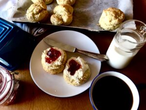 Freshly baked scones with whipped cream and jam on a white plate, accompanied by a cup of black coffee and a jug of milk. A tray of scones sits in the background on parchment paper.