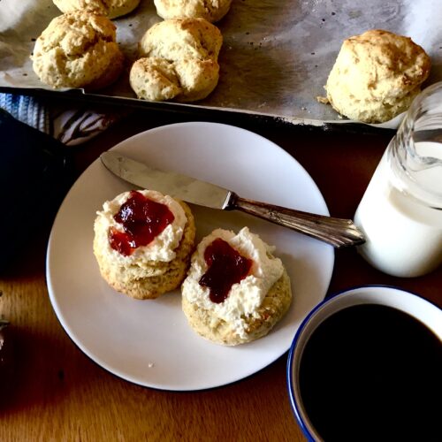 Freshly baked scones with whipped cream and jam on a white plate, accompanied by a cup of black coffee and a jug of milk. A tray of scones sits in the background on parchment paper.