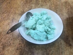 Bright blue frosting in a white bowl with a tablespoon, ready to add to the crumbs for a fun cake pop recipe for kids.