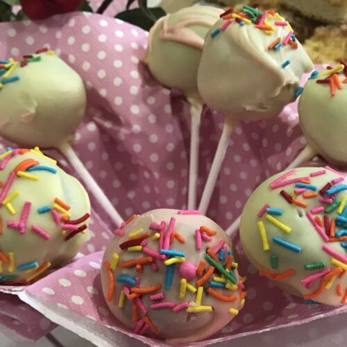 Cake pops coated in white chocolate with colourful sprinkles, arranged in a pink polka-dot paper liner.