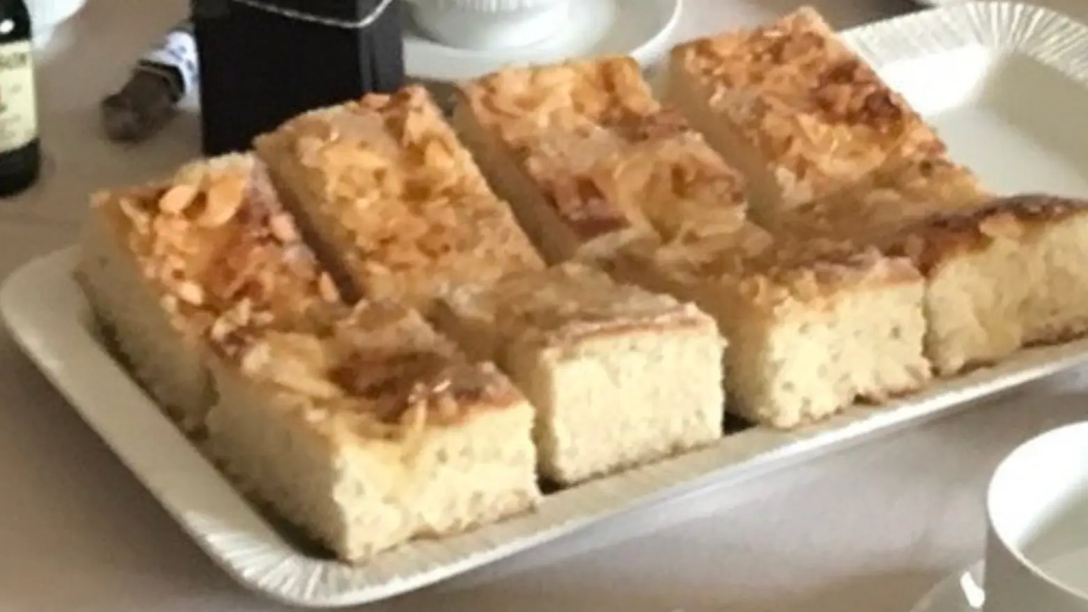 Slices of Butterkuchen, a traditional German sheet cake, with a golden crust, crunchy almond topping, and a soft, fluffy interior, served on a platter with a cup of coffee in the background.