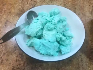 Bright blue frosting in a white bowl with a tablespoon, ready to add to the crumbs for a fun cake pop recipe for kids.