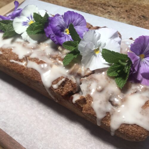 Apple Cinnamon Swirl Loaf cake with a light glaze, topped with purple and white edible flowers and fresh mint leaves, placed on a wooden board with parchment paper.