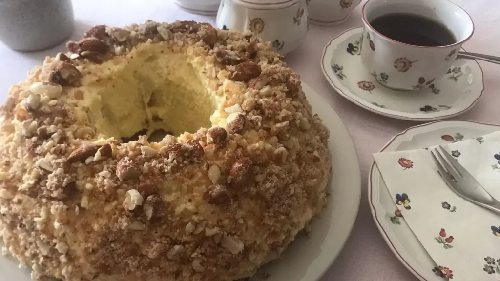 Frankfurter Kranz cake with buttercream and krokant on a white plate. A floral tea set, coffee, and napkins are arranged on a table.