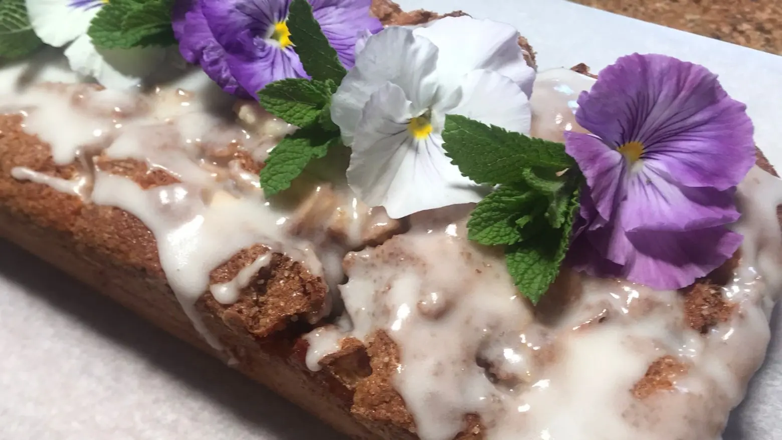 Apple Cinnamon Swirl Loaf cake with a light glaze, topped with purple and white edible flowers and fresh mint leaves, placed on a wooden board with parchment paper.