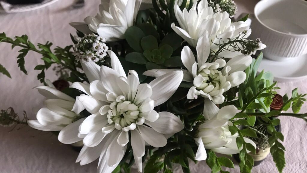 White floral arrangement with chrysanthemums, greenery, and small white blossoms in a simple table setting.