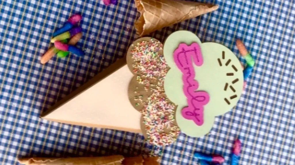 A handmade cardboard ice cream cone treat box, featuring layered scoops with sprinkles, placed on a checkered background with a waffle cone and colourful gummy candies scattered around.