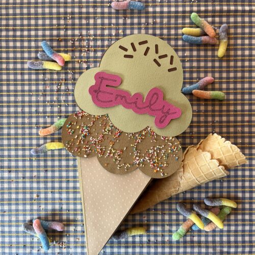A handmade cardboard ice cream cone treat box, featuring layered scoops with sprinkles, placed on a checkered background with a waffle cone and colourful gummy candies scattered around.