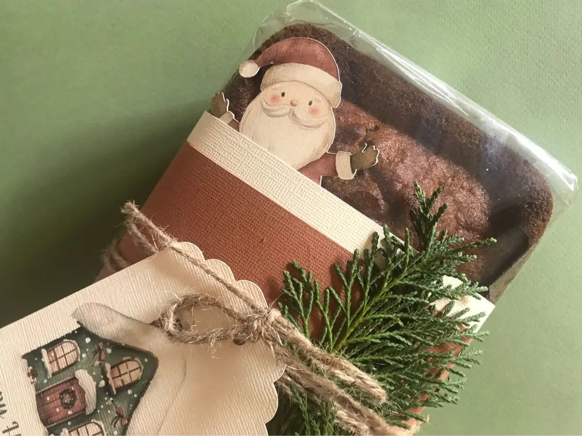 A gift-wrapped loaf of banana bread decorated with a cutout of Santa, greenery, and rustic twine on a green background.