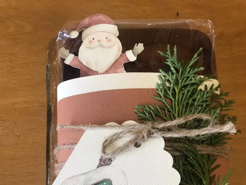 A gift-wrapped loaf of banana bread decorated with a cutout of Santa, greenery, and rustic twine and a gift tag.