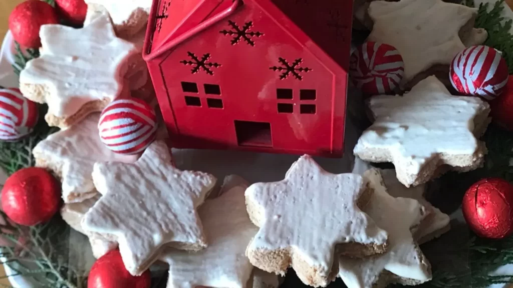 A festive plate of traditional German Zimtsterne (cinnamon star cookies) topped with white icing, surrounded by red and white foil-wrapped chocolates and evergreen sprigs, with a red house in the centre.