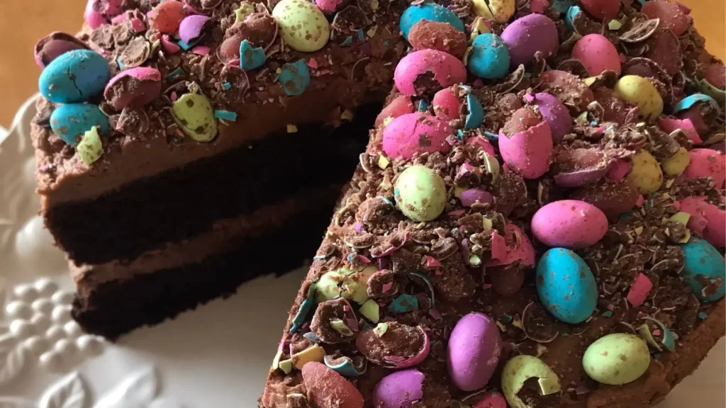 Homemade chocolate cake with creamy frosting, topped with colourful crushed Easter eggs, served on a white floral plate.