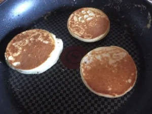 Three golden-brown crumpets cooking in a non-stick frying pan,