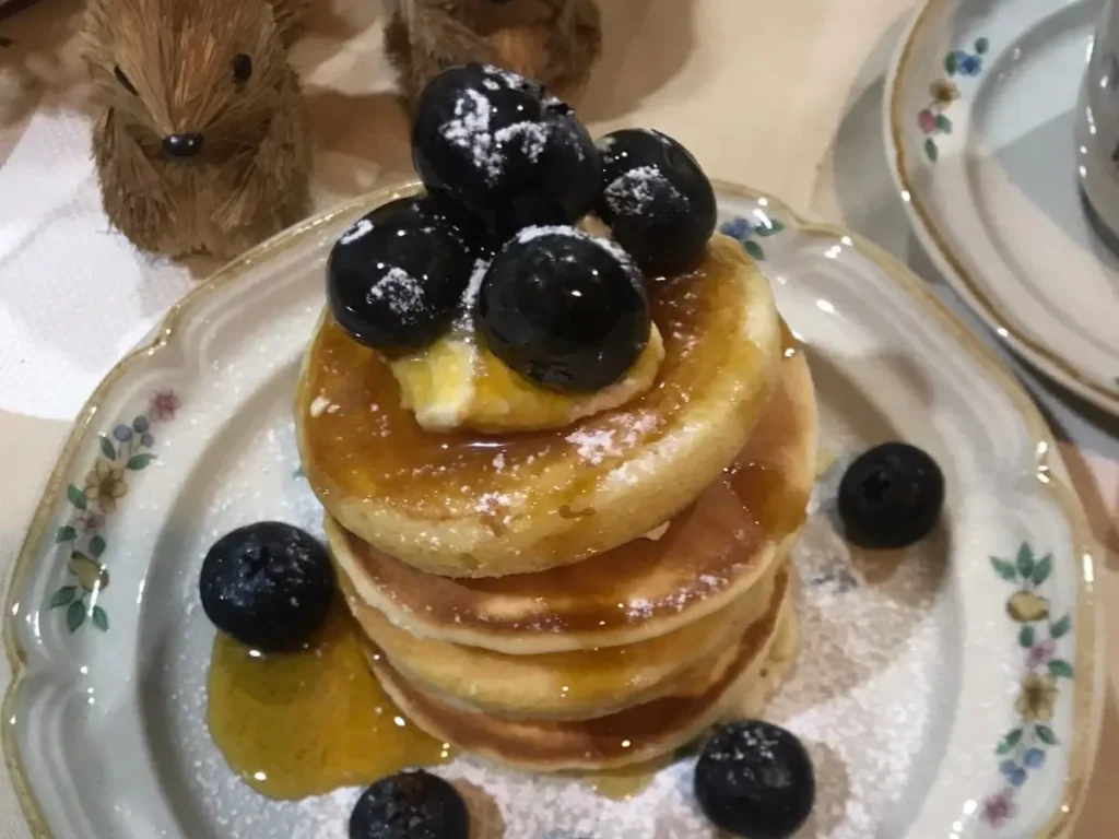 Stack of golden South African crumpets drizzled with syrup, topped with fresh blueberries and a light dusting of icing sugar, served on a floral plate.