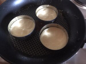 Three metal crumpet rings filled with batter sitting in a frying pan, beginning to cook on the stovetop.