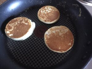 Three golden-brown crumpets cooking in a non-stick frying pan, freshly flipped to reveal their crisp, toasted surfaces.