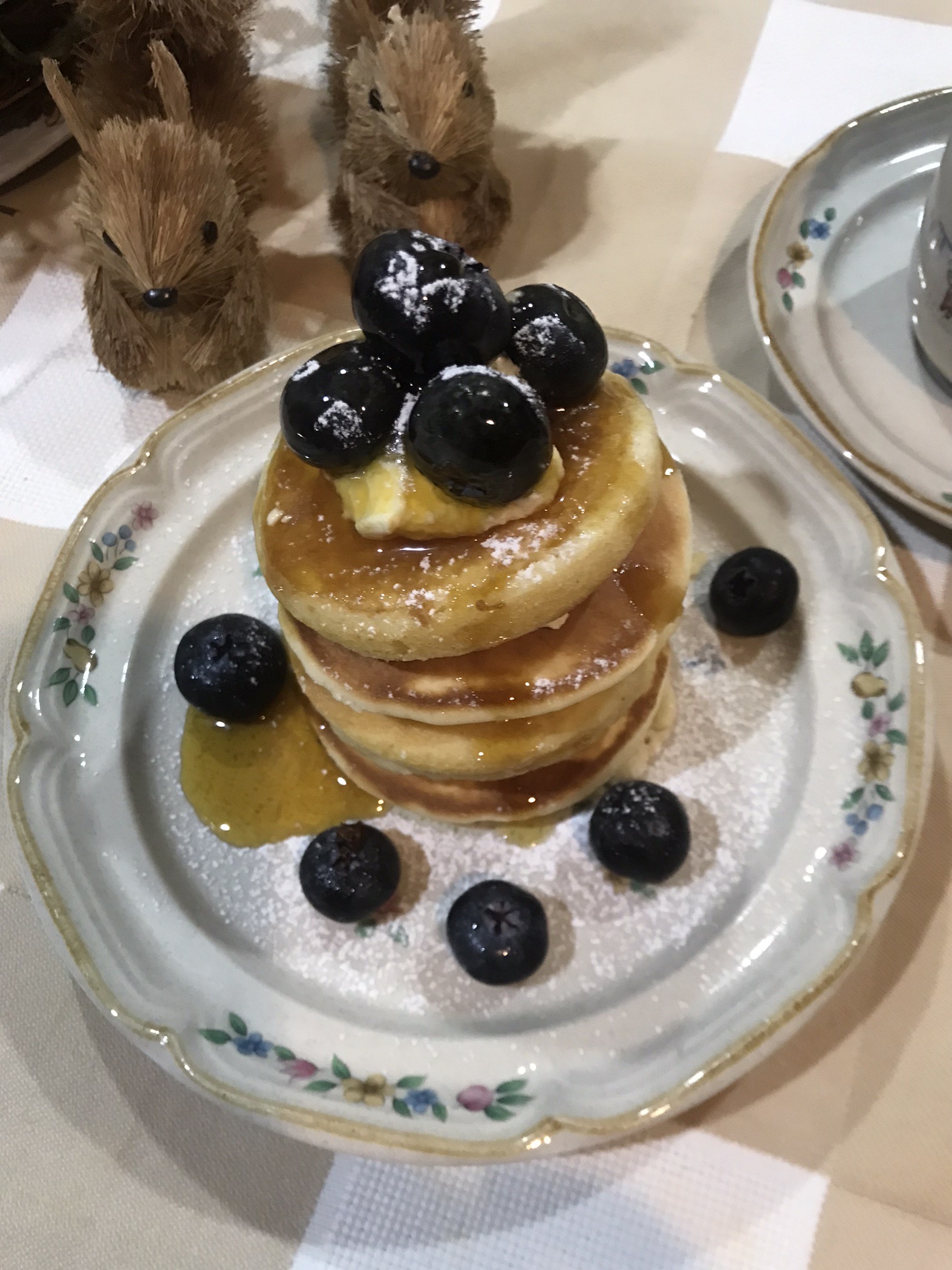 A stack of golden crumpets with whipped cream, syrup, and blueberries on a floral plate, dusted with powdered sugar.