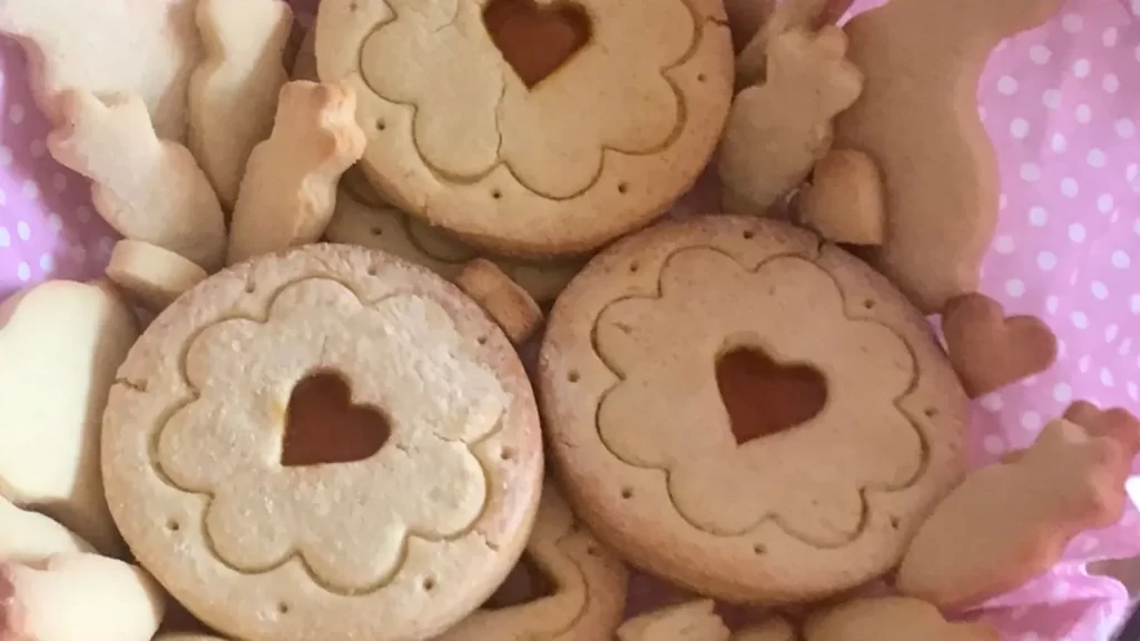 A close-up of German Tea Biscuits (Teegebäck) with heart-shaped jam-filled centers, surrounded by bunny and carrot-shaped cookies on a pink polka dot napkin.