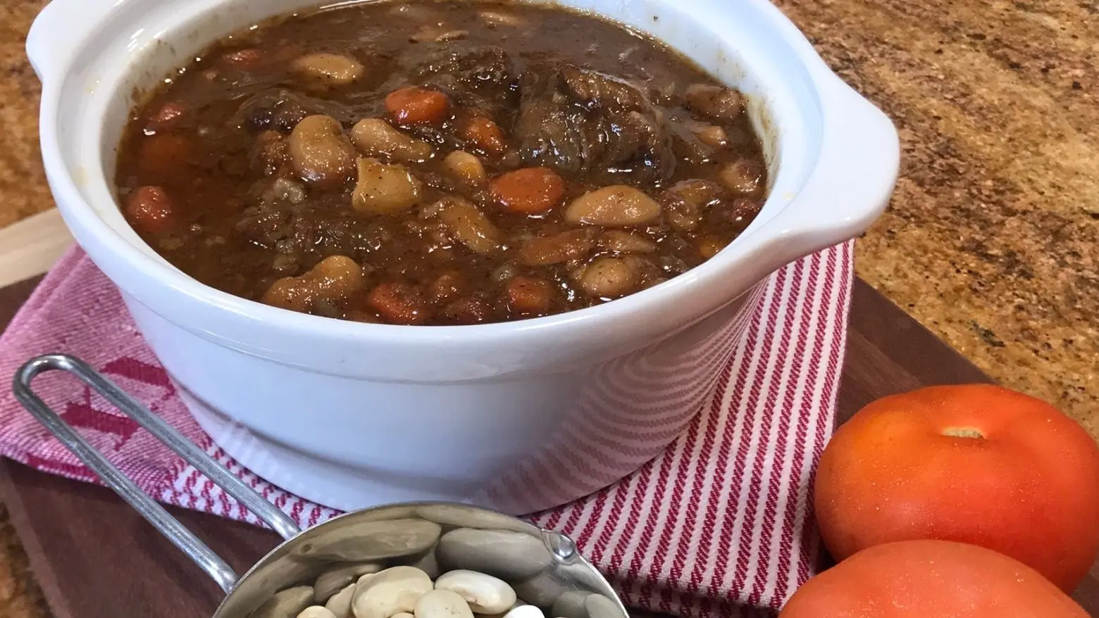 Slow-Braised Oxtail Stew Recipe - A white ceramic bowl filled with oxtail stew, featuring white beans, carrots, and a thick brown sauce. The dish is placed on a wooden board with a red-striped cloth, next to fresh tomatoes and a measuring cup of dry white beans.