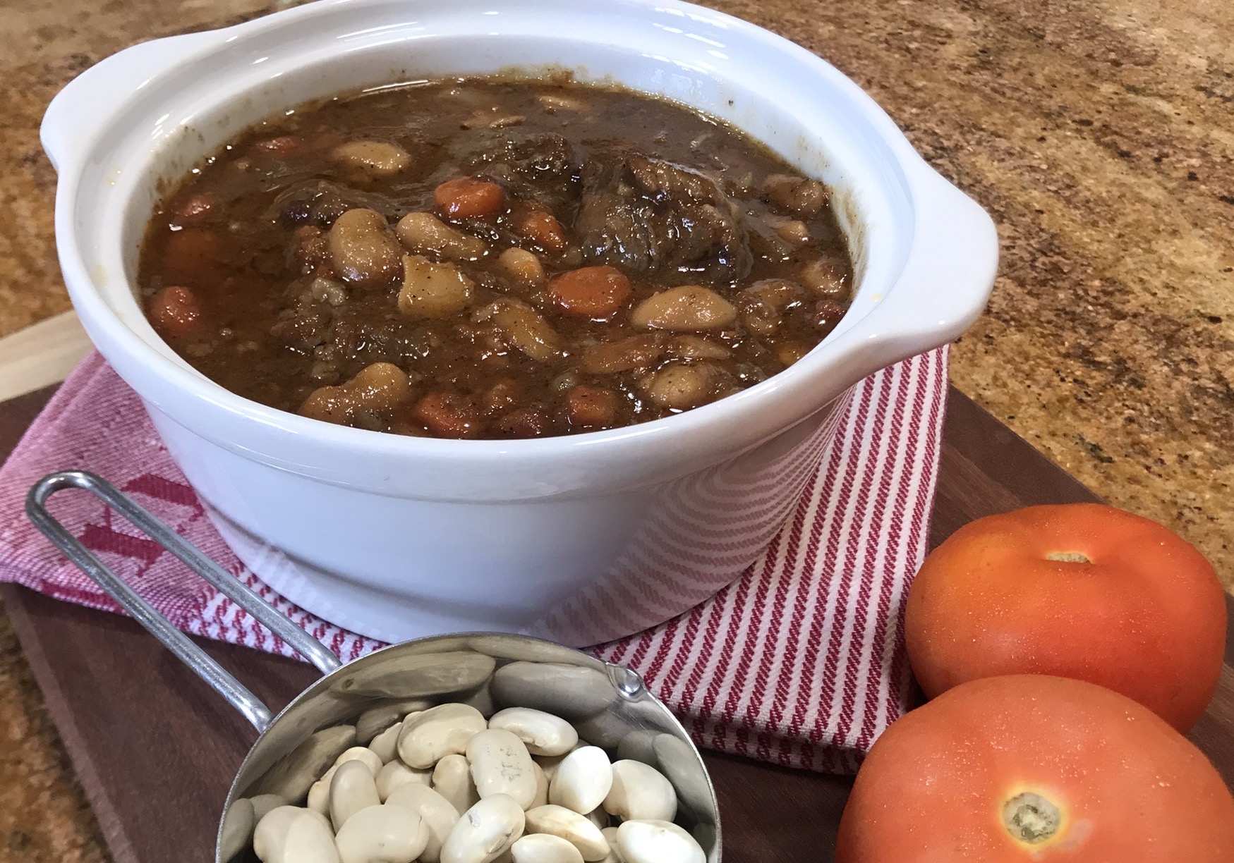 Slow-Braised Oxtail Stew Recipe - A white ceramic bowl filled with oxtail stew, featuring white beans, carrots, and a thick brown sauce. The dish is placed on a wooden board with a red-striped cloth, next to fresh tomatoes and a measuring cup of dry white beans.