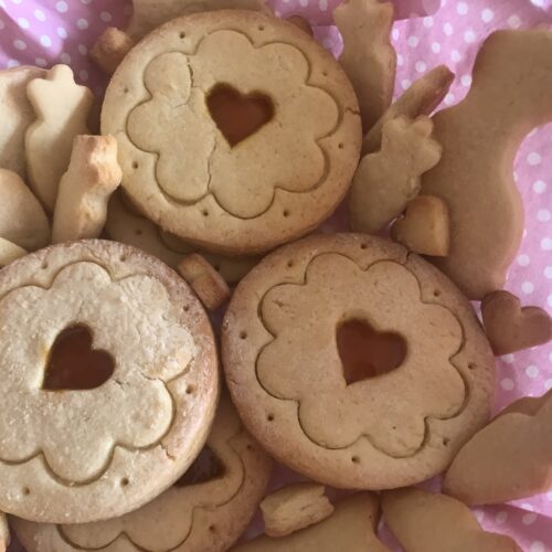A close-up of German Tea Biscuits (Teegebäck) with heart-shaped jam-filled centers, surrounded by bunny and carrot-shaped cookies on a pink polka dot napkin.