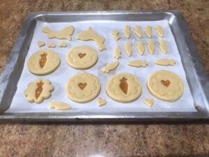 Unbaked cookies on a parchment-lined baking tray, featuring round cookies with heart and carrot cutouts filled with apricot jam, alongside small shaped cookies in carrot, rabbit and heart designs.