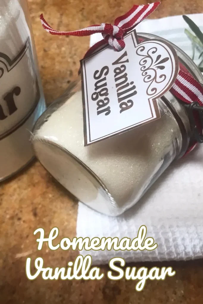 A glass jar of homemade vanilla sugar with a decorative label and red ribbon, placed on a white towel next to a sprig of thyme with a larger sugar container in the background.