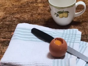 Egg opened with yolk still inside, cup ready to catch it, and a dishcloth beneath