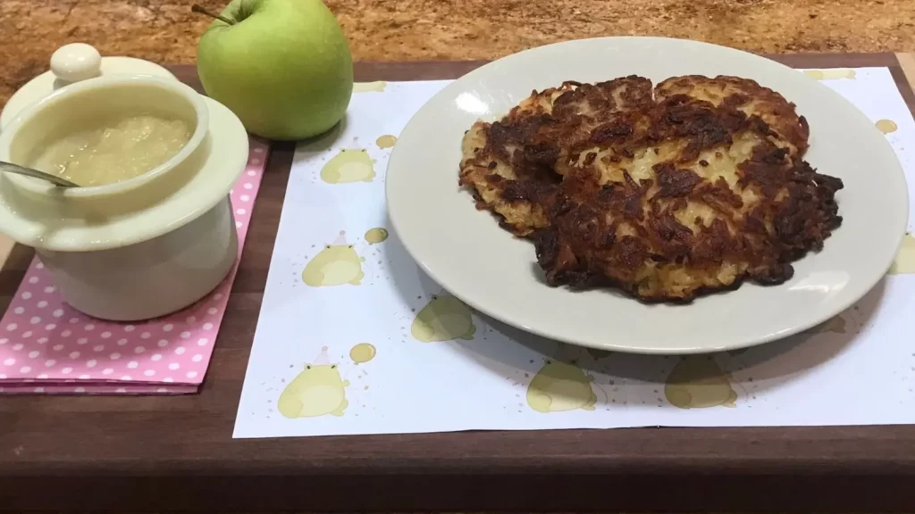 Plate of crispy German potato pancakes (Reibekuchen) served with a pot of applesauce and a fresh green apple on a wooden tray.