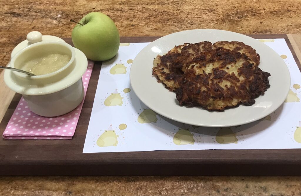 Plate of crispy German potato pancakes (Reibekuchen) served with a pot of applesauce and a fresh green apple on a wooden tray.