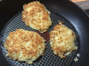 Three golden-brown Kartoffelpuffer (German potato pancakes) frying in a non-stick pan.