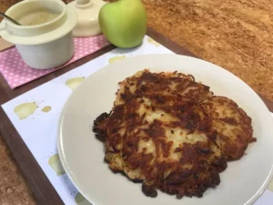 Plate of crispy German potato pancakes (Reibekuchen) served with a pot of applesauce and a fresh green apple on a wooden tray.