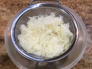 Freshly grated potatoes draining in a metal strainer over a plastic bowl on a kitchen counter.