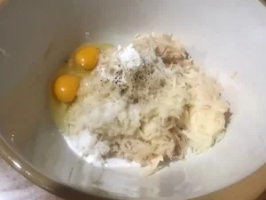 Grated potatoes with eggs, cornflour, salt, and pepper in a large mixing bowl, ready to make potato pancakes.