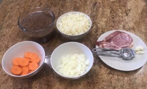 Chopped ingredients for homemade lentil soup including carrots, onions, potatoes, lentils, smoked pork, and garlic on a kitchen counter.
