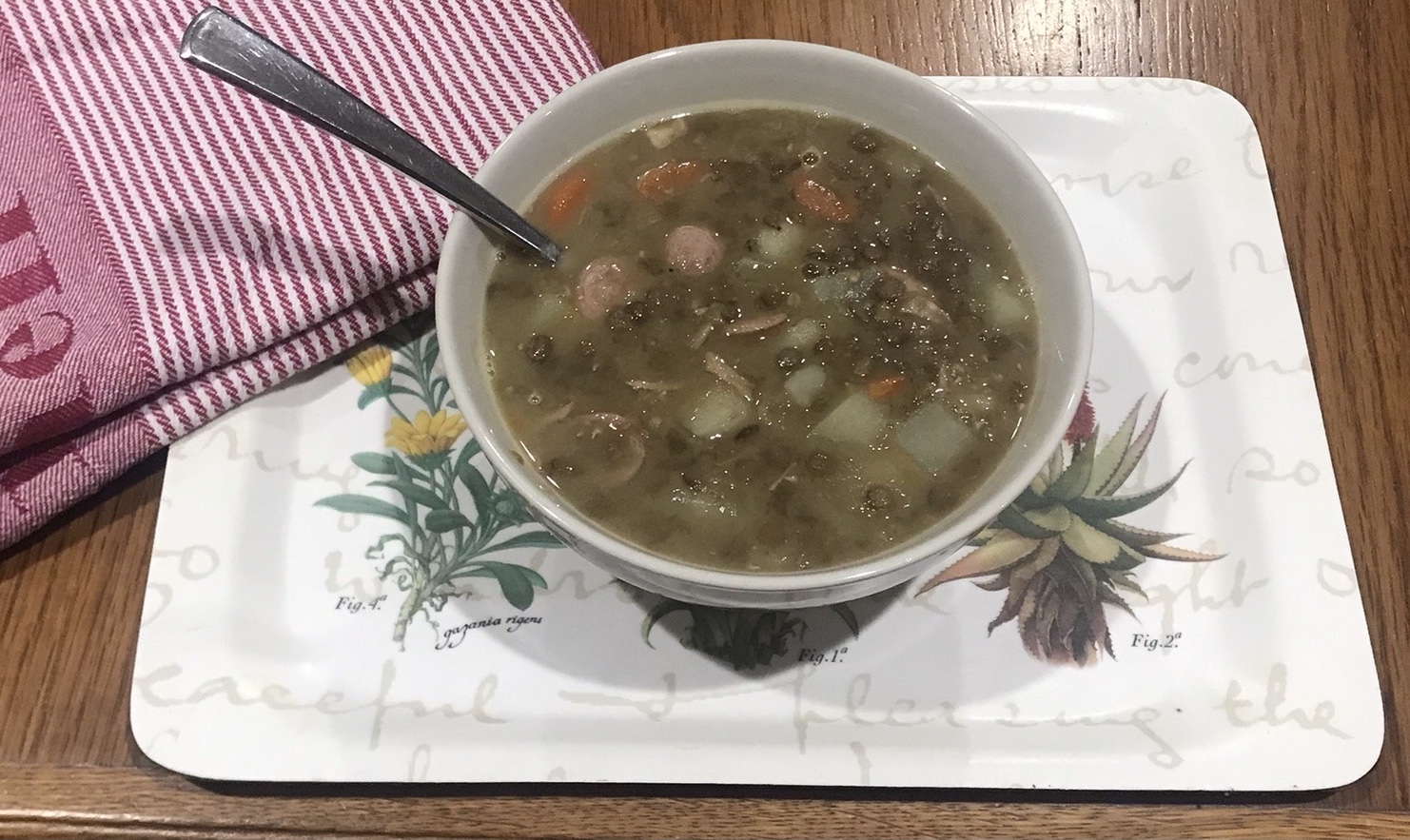 Bowl of homemade lentil soup with sausage, potatoes, and carrots on a decorative tray with a red-striped dish cloth.