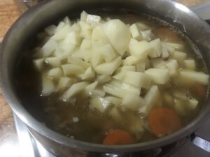 Chopped raw potatoes added to the soup pot, now filled with broth, lentils, carrots, and Kassler chops, ready to simmer into a filling homemade meal.