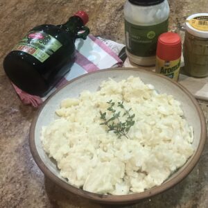 Bowl of creamy German potato salad garnished with fresh thyme, surrounded by ingredients like mayonnaise, mustard, and olive oil.
