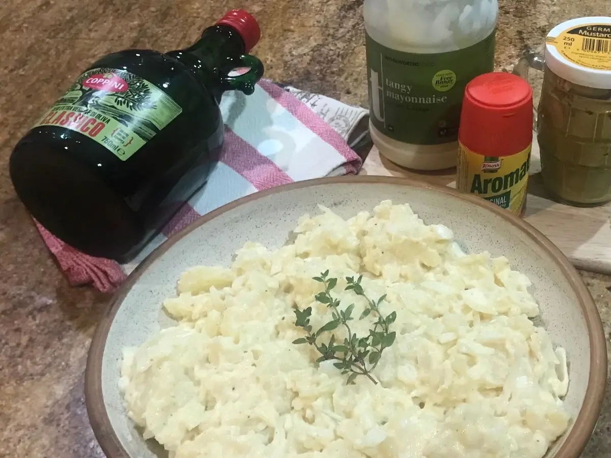 Bowl of creamy German potato salad garnished with fresh thyme, surrounded by ingredients like mayonnaise, mustard, and olive oil.