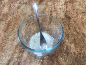 Lightly beaten egg white in a glass bowl with a fork on a brown speckled countertop.