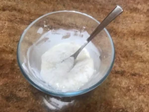 Tylose paste has been sprinkled ontop a mixture of egg white and icing sugar in a glass bowl with a fork on a brown countertop.