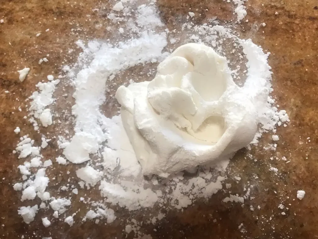 A small mound of homemade white gum paste sits on a lightly dusted surface with powdered sugar scattered around.